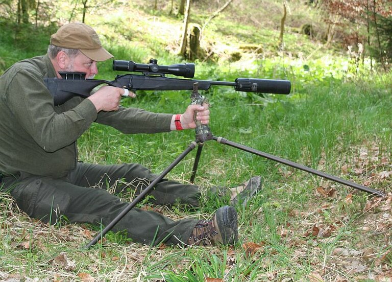 Im Wald sitzt ein Jäger auf dem Boden und zielt mit seinem Gewehr. Die Waffe liegt auf einem Dreibein.